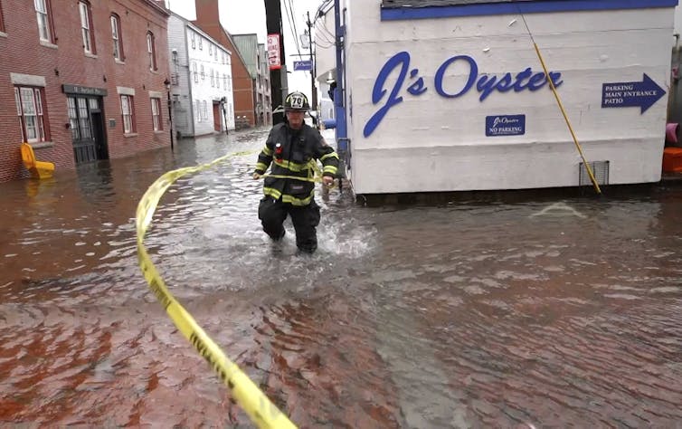 A firefighter wades through knee-deep seawater on a shopping street with a sign for J's Oysters behind him.